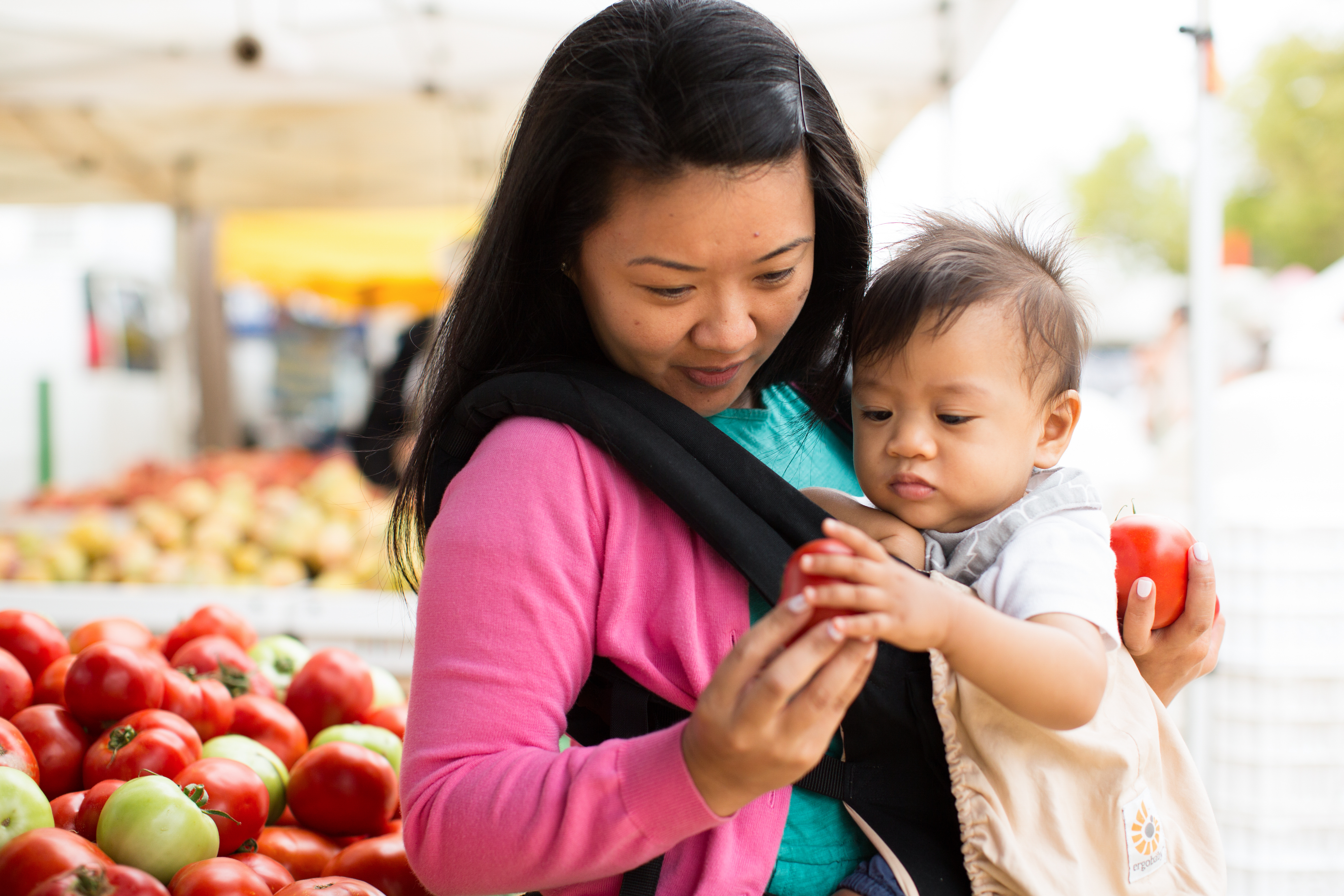 Mother Holding Child in Baby Carrier Looking at Tomatoes at the Farmer's Market